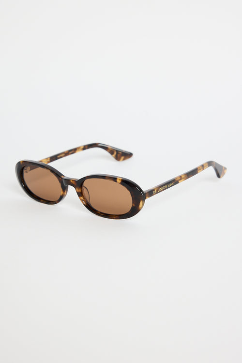 Narrow tortoiseshell oval sunglasses with warm brown tinted lenses, photographed against a white background