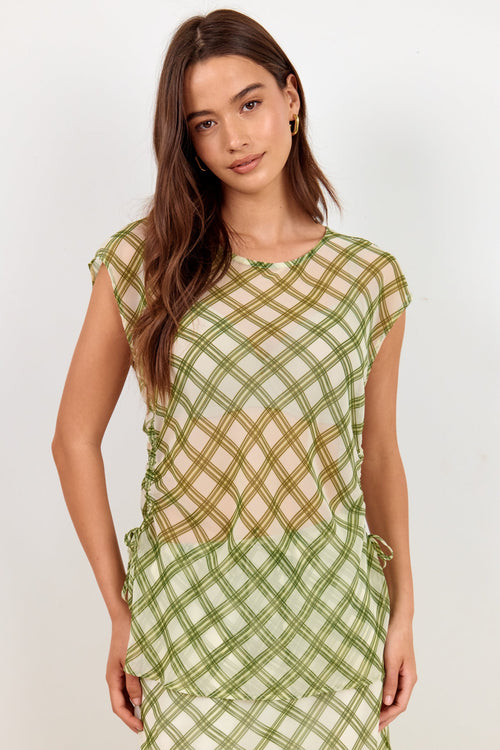 A woman modeling a sheer olive green mesh dress with a crisscross grid pattern over a light beige underlayer, set against a plain off-white studio background.