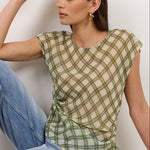 A woman sits on a white floor against a white wall wearing a green and white plaid sleeveless top with side ruffles and light blue jeans.