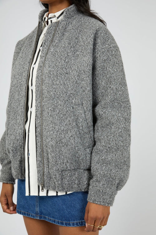 Model wears a grey marle bomber jacket with a textured wool-blend finish. The jacket features a high collar, front zip closure, and relaxed fit with side pockets. Layered over a striped button-down shirt and styled with a denim mini skirt for a casual yet elevated outfit.