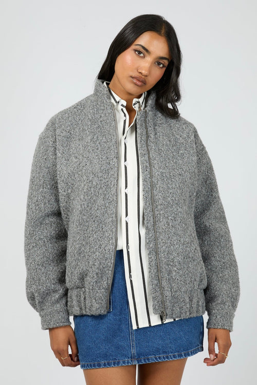 Model wears a grey marle bomber jacket with a textured wool-blend finish. The jacket features a high collar, front zip closure, and relaxed fit with side pockets. Layered over a striped button-down shirt and styled with a denim mini skirt for a casual yet elevated outfit.