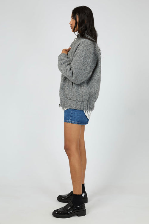 Model wears a grey marle bomber jacket with a textured wool-blend finish. The jacket features a high collar, front zip closure, and relaxed fit with side pockets. Layered over a striped button-down shirt and styled with a denim mini skirt for a casual yet elevated outfit.