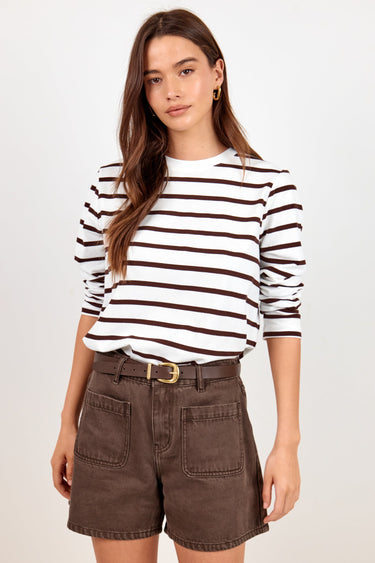 A woman modeling a white long-sleeve knit top with dark brown horizontal stripes, tucked into dark brown denim shorts with a matching belt, against a plain light gray studio background.