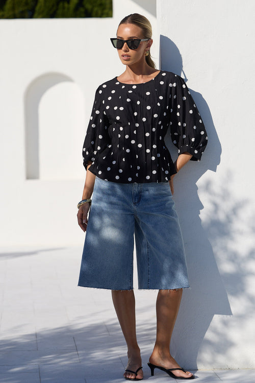 A woman stands outdoors against a white wall wearing a black long-sleeve blouse with white polka dots, light blue denim culottes, and black sandals.