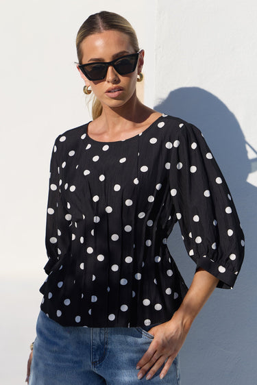 A woman wearing a black long-sleeve blouse with white polka dots and blue jeans, standing against a white wall in bright sunlight.