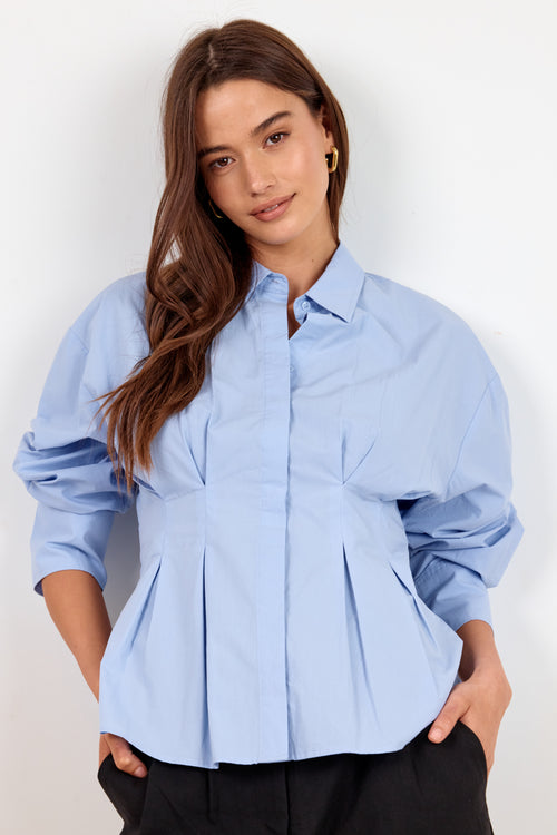 A woman wearing a light blue button-up blouse with dramatic puffed sleeves and a gathered peplum waist, paired with black pants, against a plain white background.