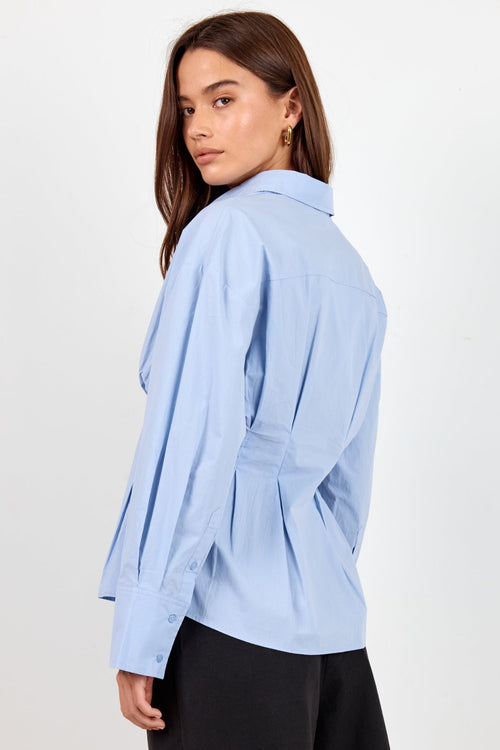 A woman modeling a light blue long-sleeve button-up shirt with a belted waist, paired with black shorts, photographed against a plain off-white studio background.