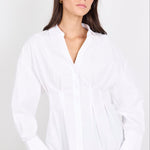 A woman wearing a white long-sleeve button-up blouse with a gathered waist and peplum-like drape, paired with blue jeans, against a plain light gray studio background.