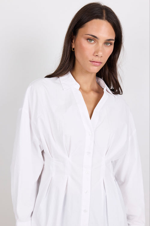 A woman wearing a white long-sleeve button-up shirt dress with a tie waist, photographed against a light gray studio background.