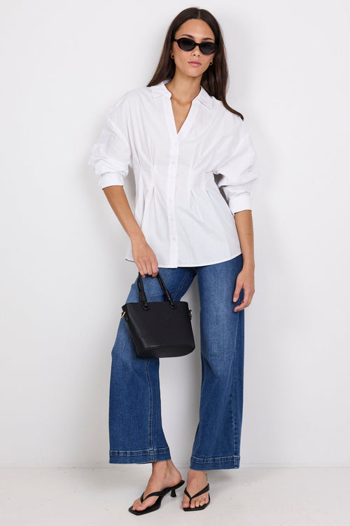 A woman modeling a white oversized button-up shirt with blue wide-leg jeans, black sandals, and a black handbag against a plain white studio background.