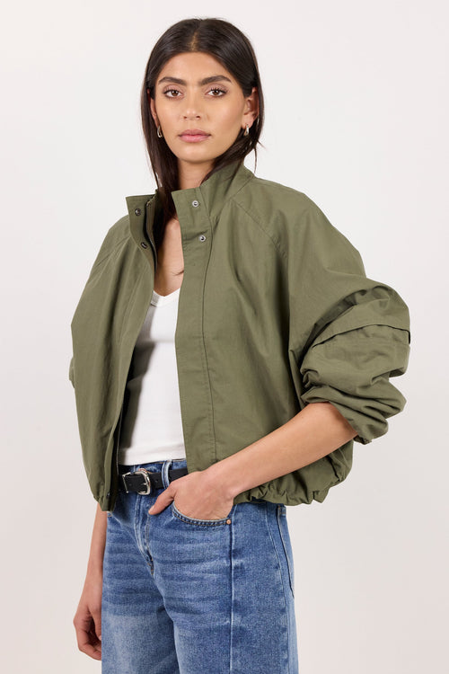 A woman modeling an olive green cropped utility jacket with voluminous rolled sleeves, worn over a white top and blue jeans, against a plain light background.