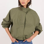 A woman modeling an olive green lightweight jacket with a high stand-up collar and voluminous sleeves, paired with blue jeans, against a plain light background.