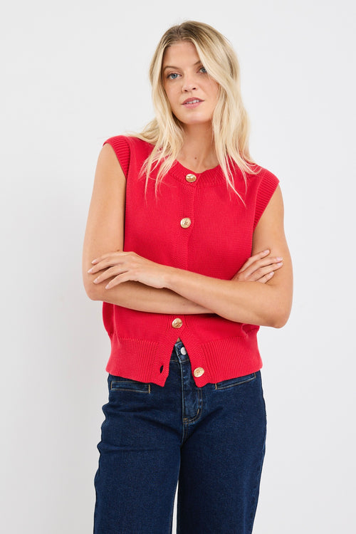 Woman wearing a sleeveless red knit vest with gold buttons down the front, styled with dark denim and loose blonde waves.