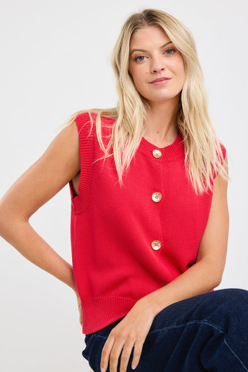 Woman wearing a sleeveless red knit vest with gold buttons down the front, styled with dark denim and loose blonde waves.