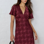Woman wearing a burgundy patterned dress against a white brick wall.