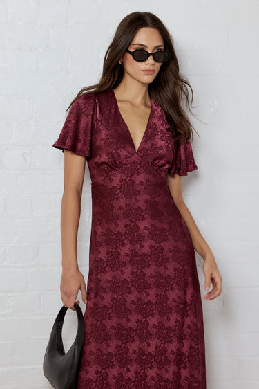Woman wearing a burgundy patterned dress against a white brick wall.