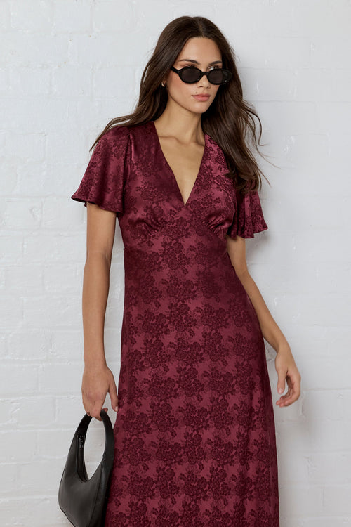Woman wearing a burgundy patterned dress against a white brick wall.