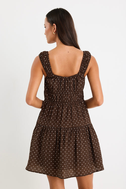 Model wears a sleeveless brown mini dress with white polka dots, featuring a tiered skirt, ruffled straps, and a tie-front detail. 