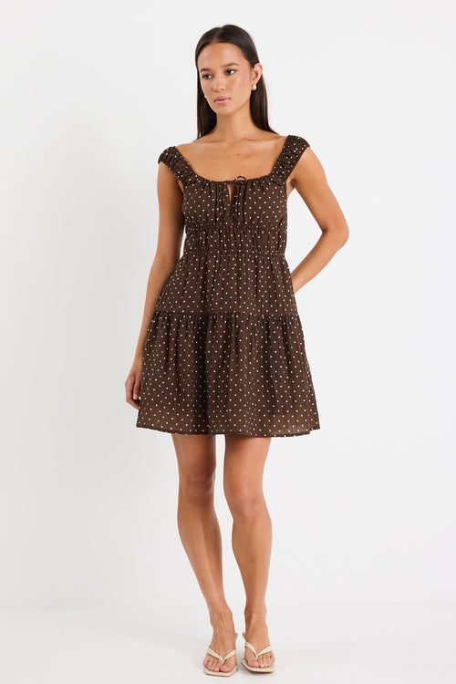 Model wears a sleeveless brown mini dress with white polka dots, featuring a tiered skirt, ruffled straps, and a tie-front detail. 