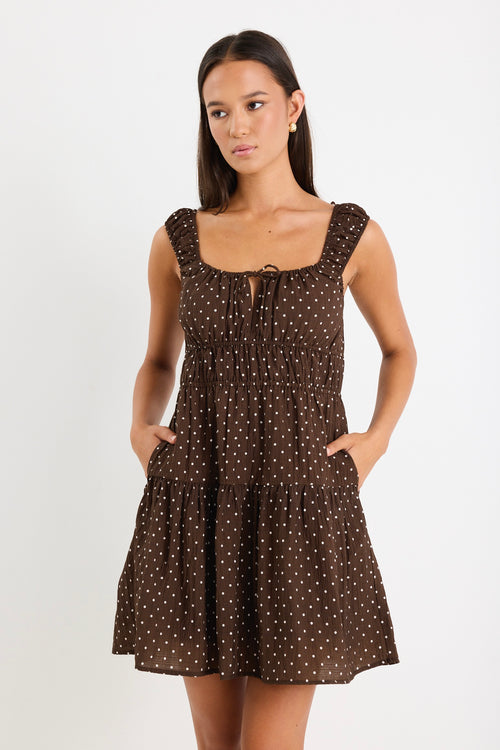 Model wears a sleeveless brown mini dress with white polka dots, featuring a tiered skirt, ruffled straps, and a tie-front detail. 