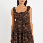 Model wears a sleeveless brown mini dress with white polka dots, featuring a tiered skirt, ruffled straps, and a tie-front detail. 