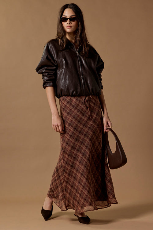 A model wearing a black leather bomber jacket, a brown and red plaid midi skirt, black pointed-toe heels, and carrying a dark brown handbag against a solid beige studio backdrop.