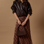 A model stands against a solid warm beige studio backdrop wearing a black leather jacket, a brown and red plaid midi skirt, black pointed flats, and holding a dark brown handbag.