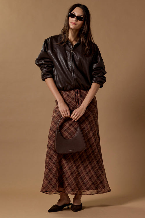 A model stands against a solid warm beige studio backdrop wearing a black leather jacket, a brown and red plaid midi skirt, black pointed flats, and holding a dark brown handbag.