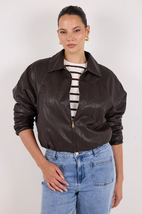 A model wearing a dark brown leather jacket with a zippered front, styled over a black and white striped t-shirt and light blue jeans, against a plain light gray background.