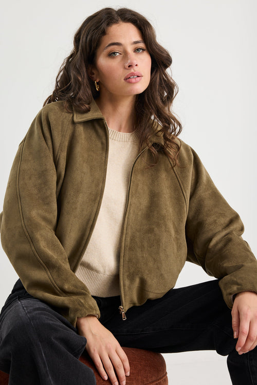Woman wearing a khaki mock suede zip-up jacket with a soft, textured finish and a relaxed fit, styled over a beige knit top and black jeans.