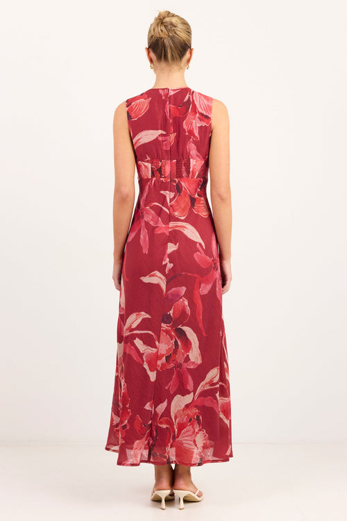 A woman viewed from behind wearing a sleeveless red maxi dress with a large pink and white floral print, standing against a plain white studio background.