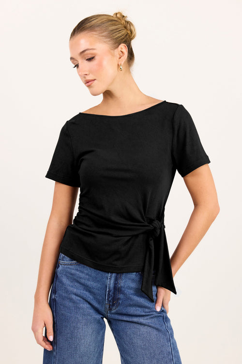 A woman wearing a black short-sleeve boat-neck top with a side tie detail, paired with blue jeans, photographed against a plain light background.