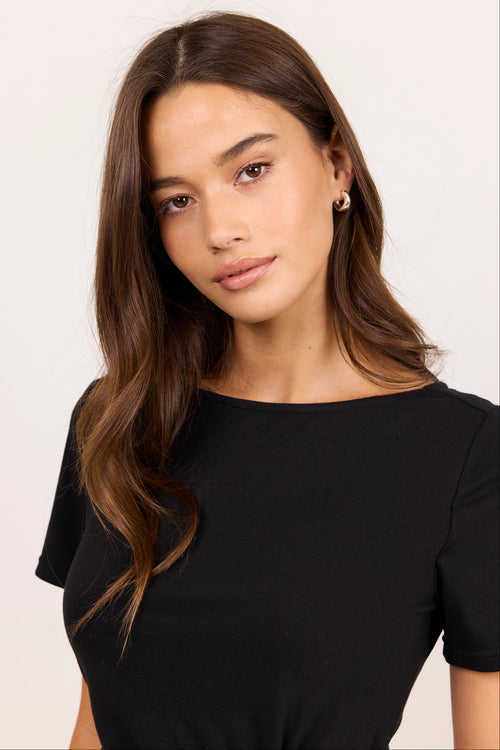 A woman with long brown hair wearing a black short-sleeve boat-neck top and small gold hoop earrings, photographed against a plain light background.