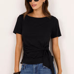 A woman modeling a black short-sleeve boat-neck t-shirt with a twisted front detail, paired with blue jeans, against a plain off-white background.