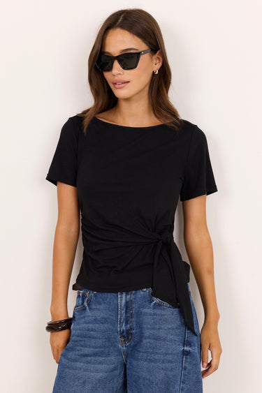 A woman modeling a black short-sleeve boat-neck t-shirt with a twisted front detail, paired with blue jeans, against a plain off-white background.