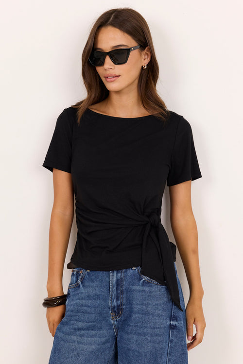 A woman modeling a black short-sleeve boat-neck t-shirt with a twisted front detail, paired with blue jeans, against a plain off-white background.