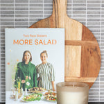 The cookbook 'More Salad' by Two Raw Sisters displayed on a dark countertop, styled with a wooden paddle board and a lit candle in front of a gray tiled wall.
