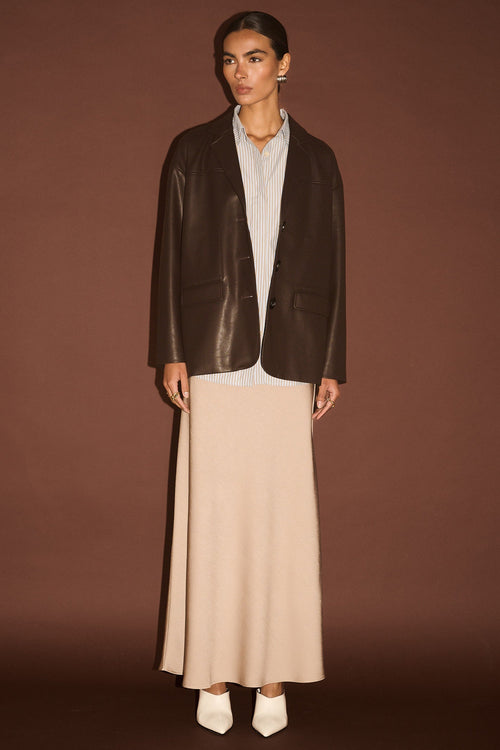 Person wearing a dark brown leather jacket over a white shirt and beige skirt on a brown background