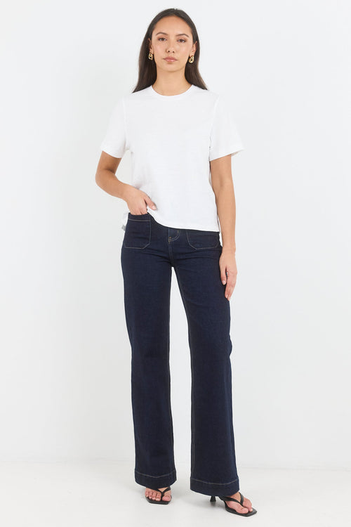 A woman modeling dark wash flare jeans with a white short-sleeve crop top and black sandals, standing against a plain white studio background.