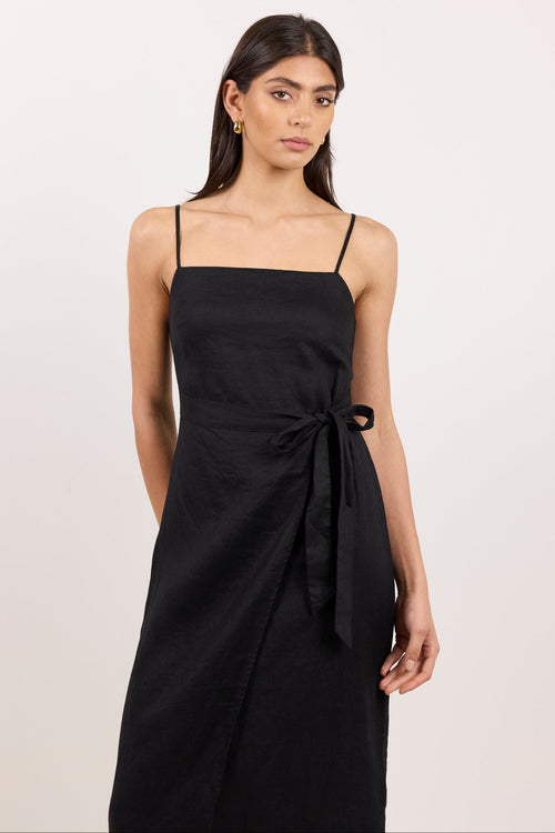 A woman modeling a black midi wrap dress with thin spaghetti straps, a square neckline, and a self-tie waist sash, photographed against a plain off-white studio background.