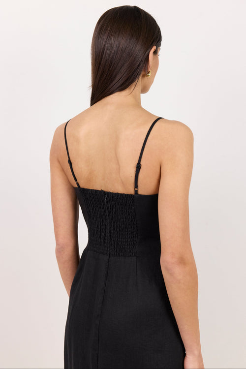 A woman viewed from the back wearing a black spaghetti-strap dress with a smocked bodice, set against a plain off-white studio background.