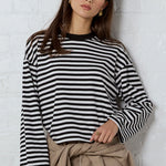 A woman stands against a white brick wall wearing a black and white striped long-sleeve top, beige cropped pants with a matching tie belt, and black flat shoes.
