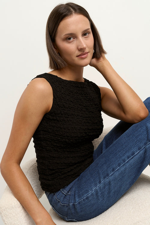 Woman wearing a black sleeveless textured top with a fitted silhouette and high neckline, paired with high-waisted denim jeans.