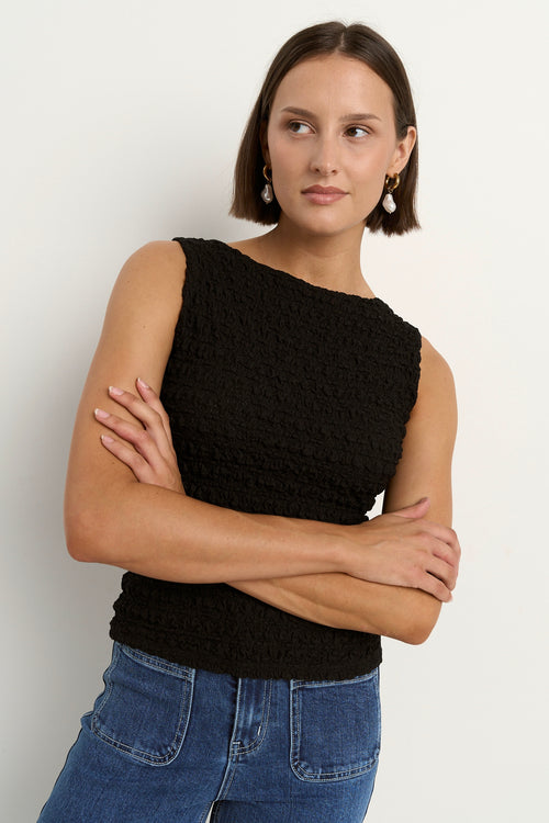 Woman wearing a black sleeveless textured top with a fitted silhouette and high neckline, paired with high-waisted denim jeans.