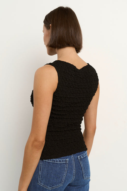 Woman wearing a black sleeveless textured top with a fitted silhouette and high neckline, paired with high-waisted denim jeans.