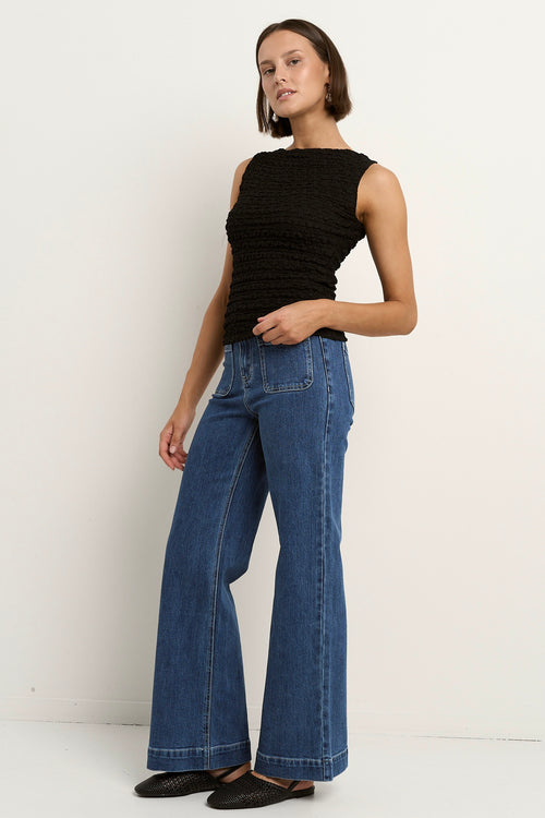 Woman wearing a black sleeveless textured top with a fitted silhouette and high neckline, paired with high-waisted denim jeans.