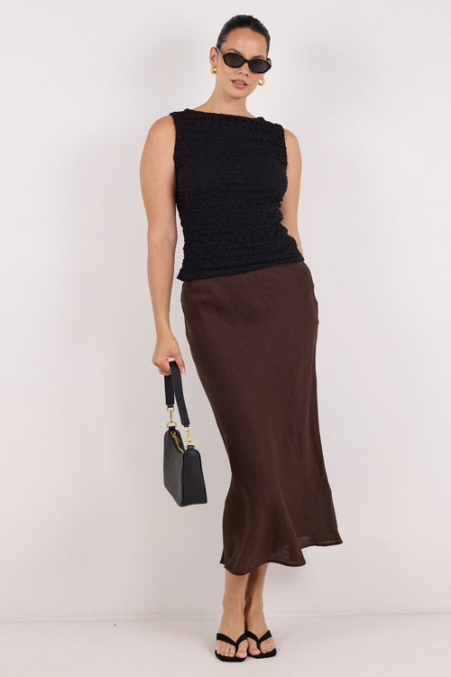 A woman modeling a sleeveless black top and a dark brown midi skirt, holding a small black handbag against a plain white studio background.