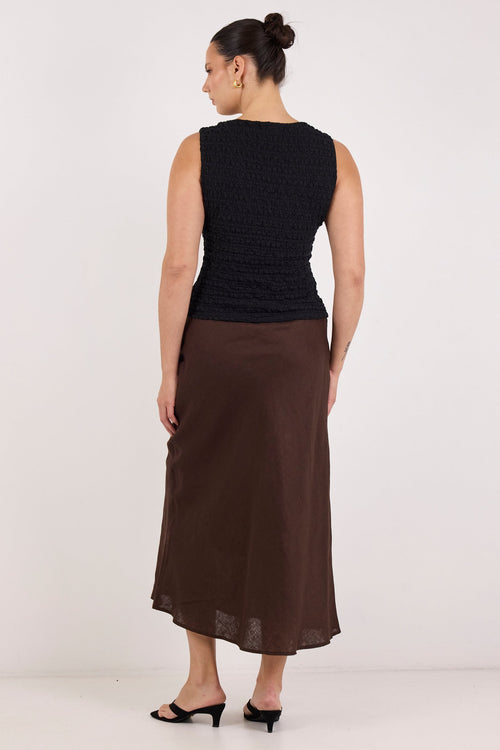 A woman is shown from the back wearing a sleeveless black textured top and a brown midi skirt, paired with black flat sandals, against a plain white studio background.
