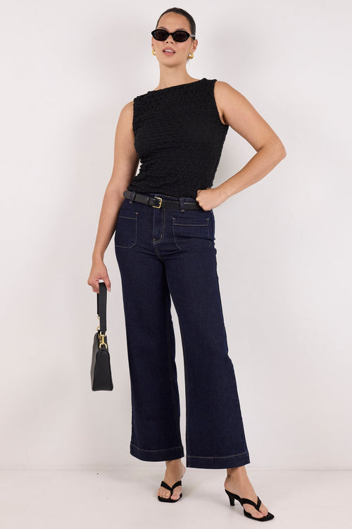A model stands against a plain white studio background wearing a black sleeveless top, dark blue wide-leg jeans with a belt, black sandals, sunglasses, and holding a small black handbag.