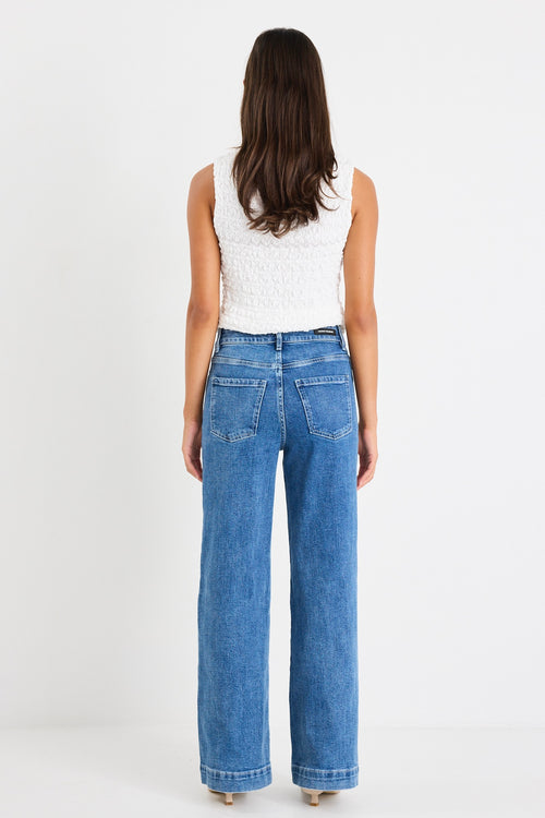 Woman wearing a white sleeveless textured top with a fitted silhouette and high neckline, paired with high-waisted denim jeans.